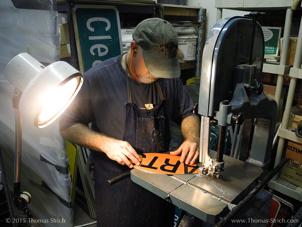 Thomas Strich stands at a bandsaw, trimming a recycled street sign so that the text reads "art"